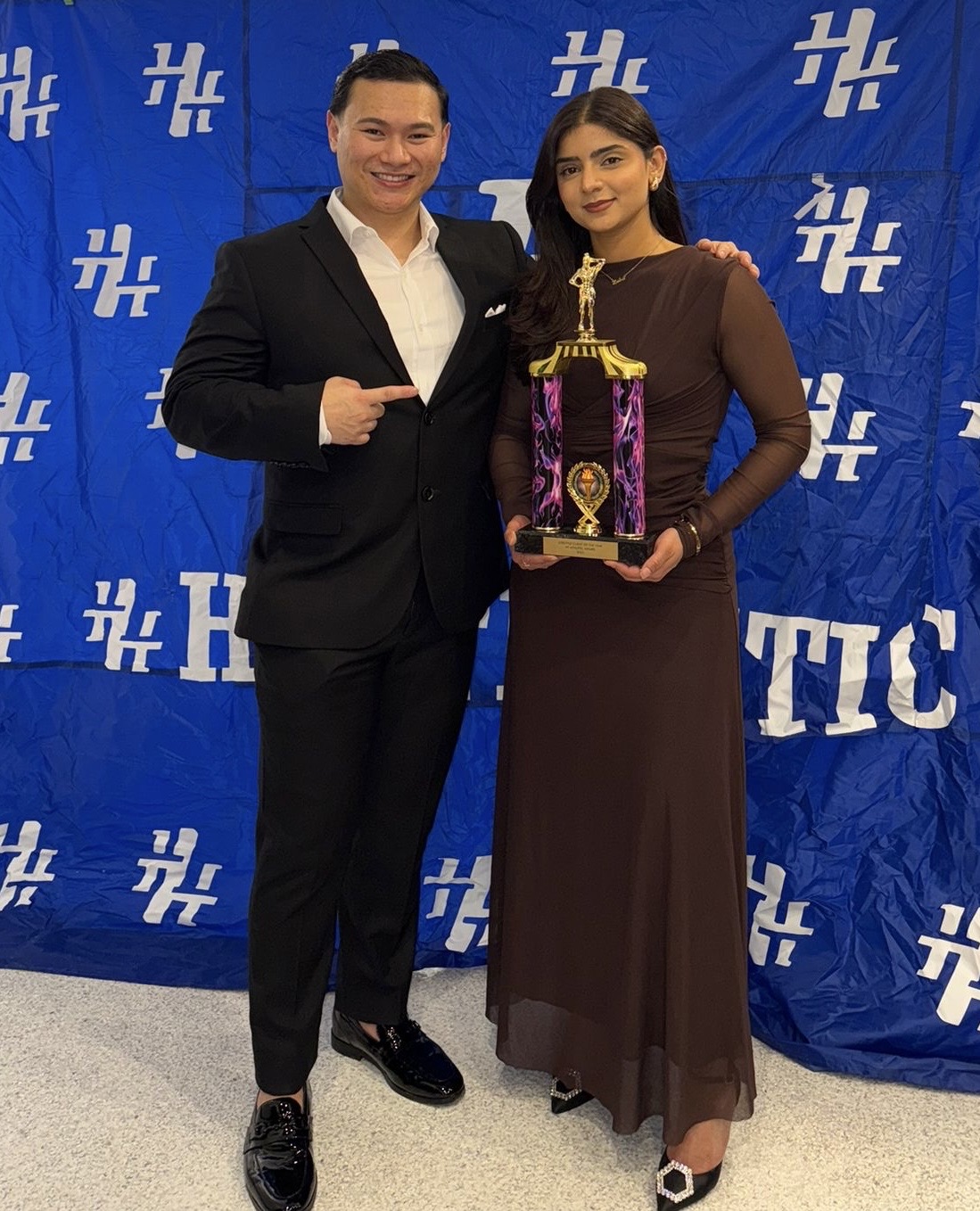 Two guests posing with a trophy in front of an HT Athletic backdrop